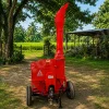 Red portable wood chipper on wheels in a rural yard with green fields and trees behind it.
